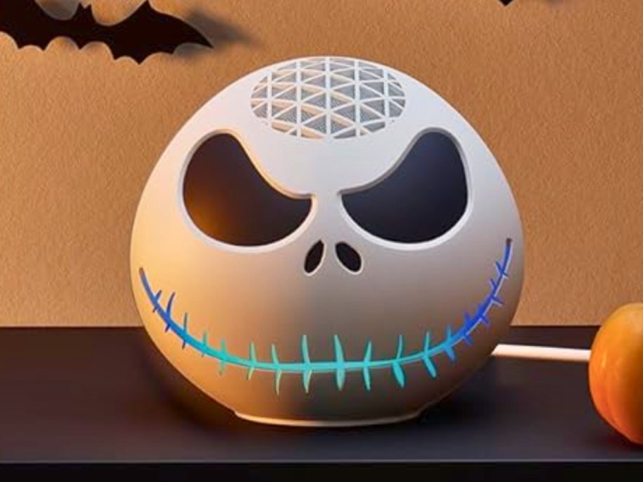 an Echo dot machine with a Jack Skellington from Nightmare Before Christmas cover sitting on a bookcase with Halloween decorative bats behind it and a faux pumpkins beside it