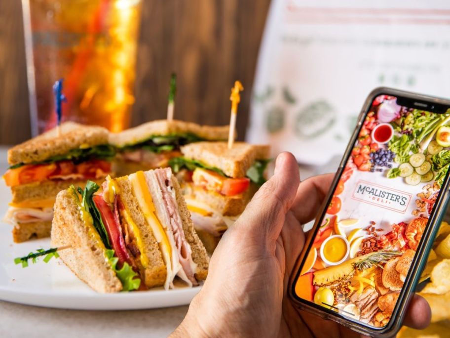 Hand holding a phone with McAlister's Rewards progra, pulled up on screen with a club sandwich and sweet tea in the background