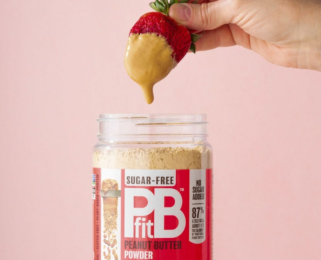 A strawberry dipped in Peanut butter over a jar of PBfit Peanut Butter Powder