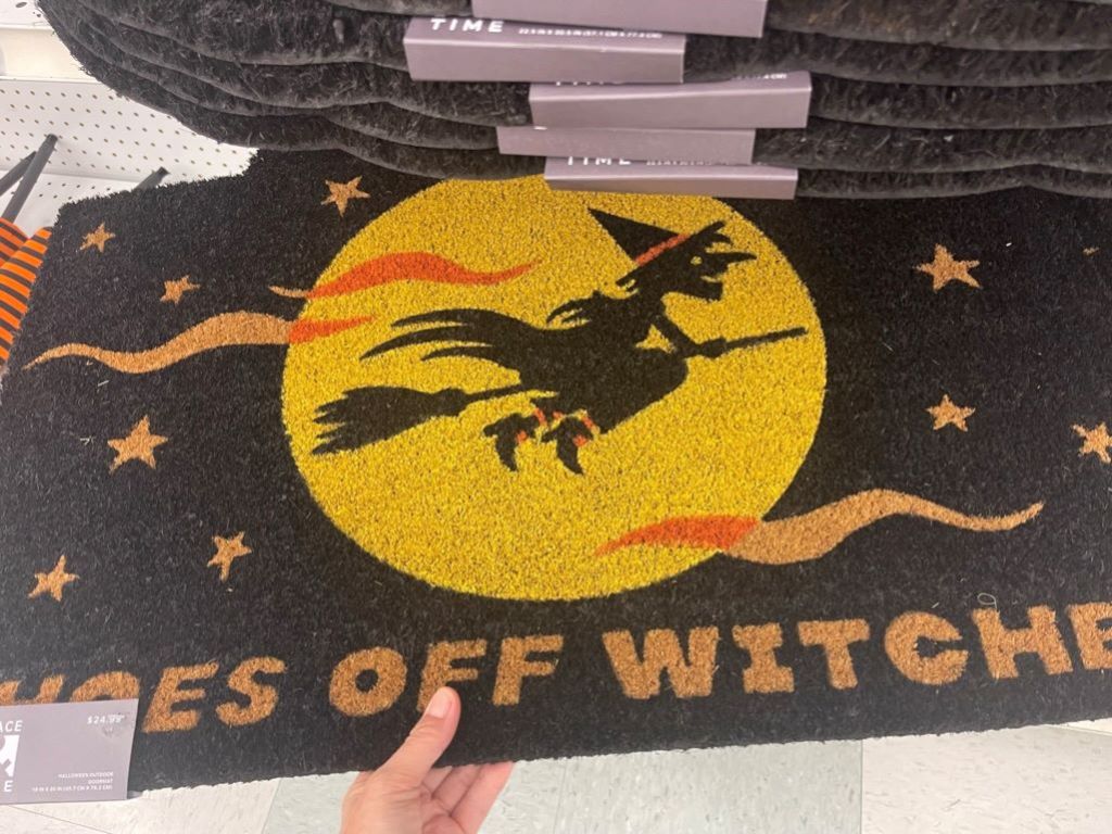 Hand holding a doormat that says shoes off witches