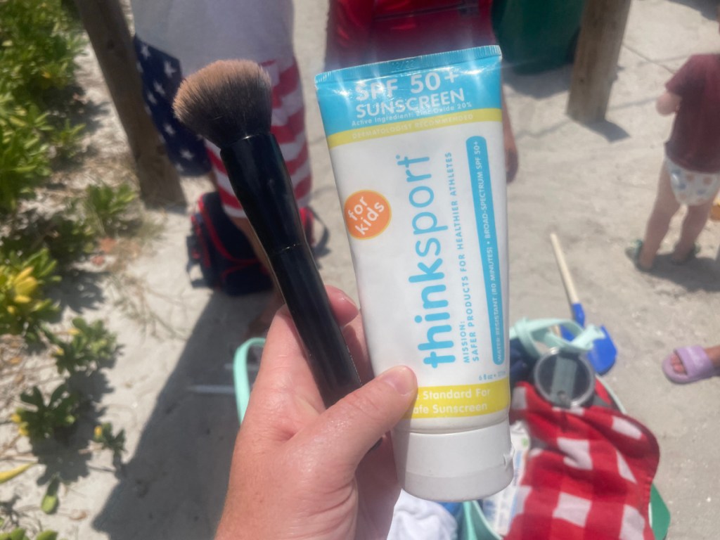 hand holding a bottle of sunscreen and a makeup brush
