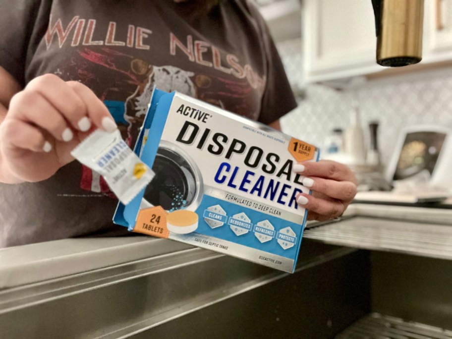 person standing by a sink holding a box of Active Appliance Cleaners for the Garbage Disposable in one hand and a pack with a single tablet in a other