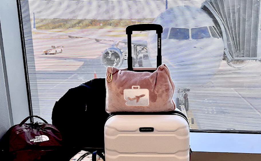 pink airplane pillow on top of suitcase with luggage in front of window with airplane