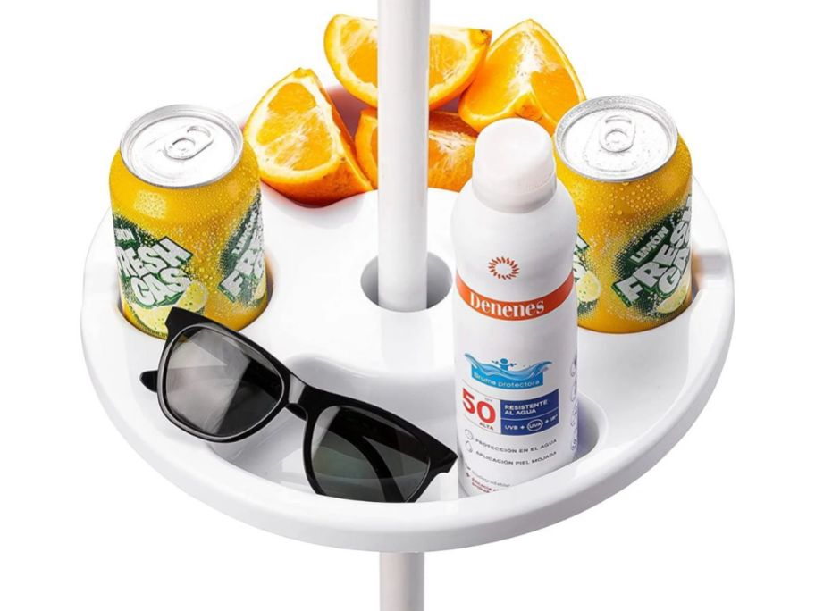 a white beach umbrella tray table with sunglasses, sun screen, and sliced oranges in the large compartments and two cans of soda in the cup holders. on a white back ground