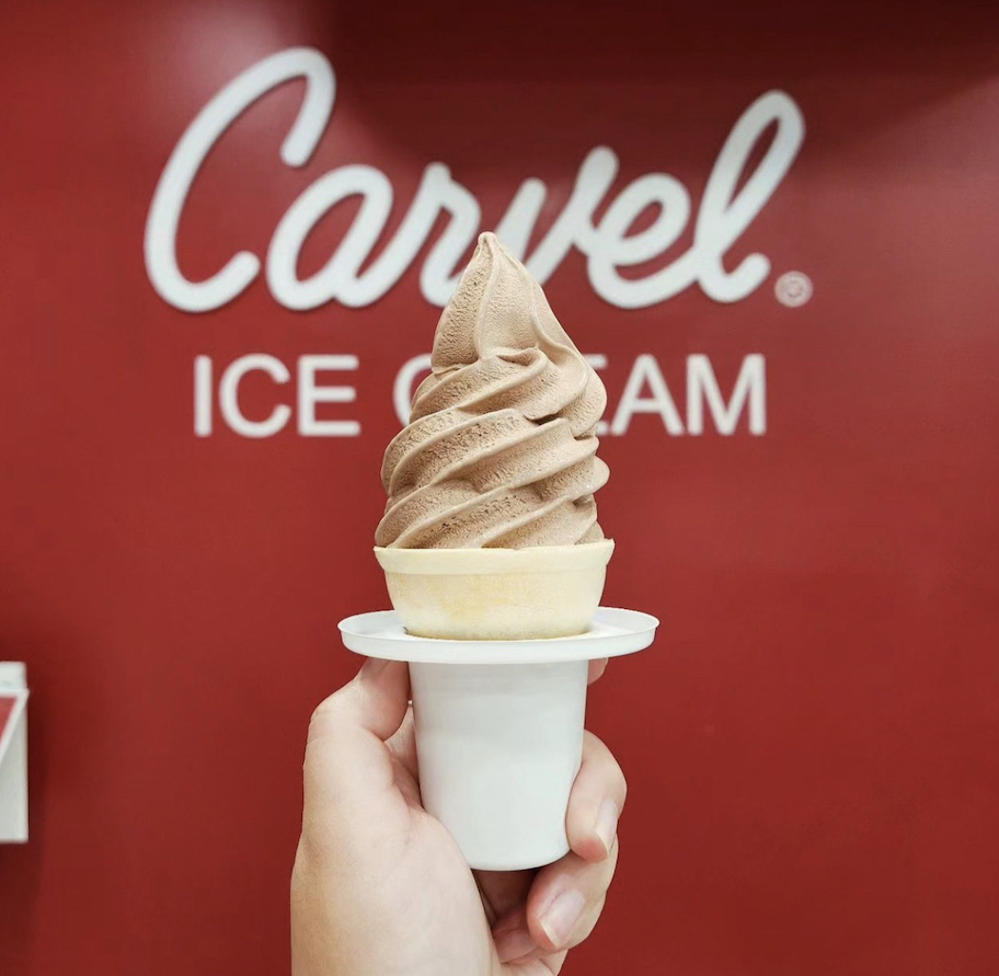 hand holding a soft serve ice cream cone in front of carvel ice cream sign