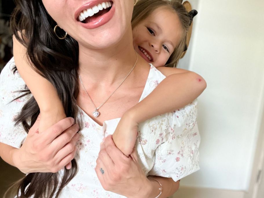 smiling woman wearing a heart shaped pendant necklace, with her daughter hugging her around the neck from behind