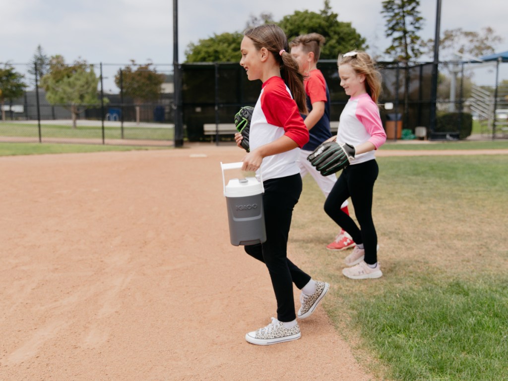 girls in softball uniforms carrying an igloo beverage jug