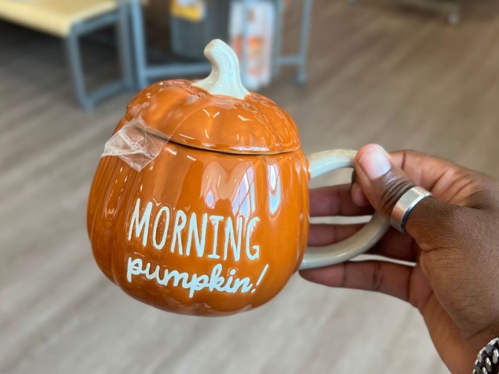 a woman's hand holding an orange pumpkin shaped coffee mug with morning pumpkin painted on one side from kohls