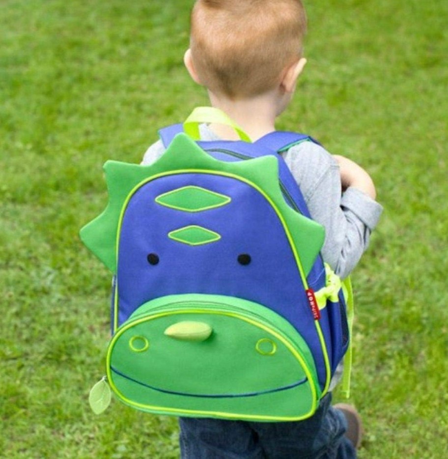 little boy walking in grass wearing blue and green dinosaur backpack