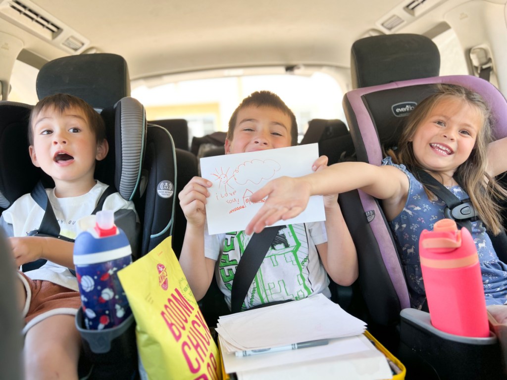 three kids having fun and joking around for the camera while buckled into their carseats