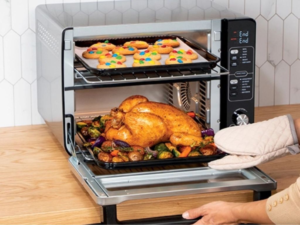Ninja 12-in-1 Double Oven with FlexDoor shown with woman's hand in oven mitt taking roasted chicken out of the bottom and cookies on the top