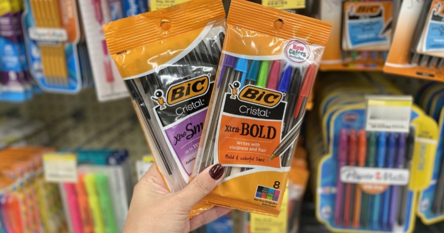 hand holding 2 packs of Bic Cristal pens, with more Bic pens and highlighters in the background