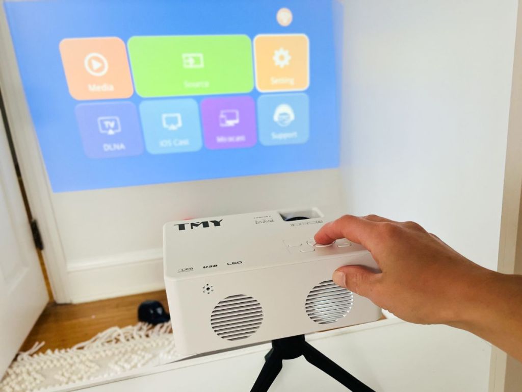 TMY Mini Projector with Tripod shown in living room with screen projecting on wall and woman's hand on the projector