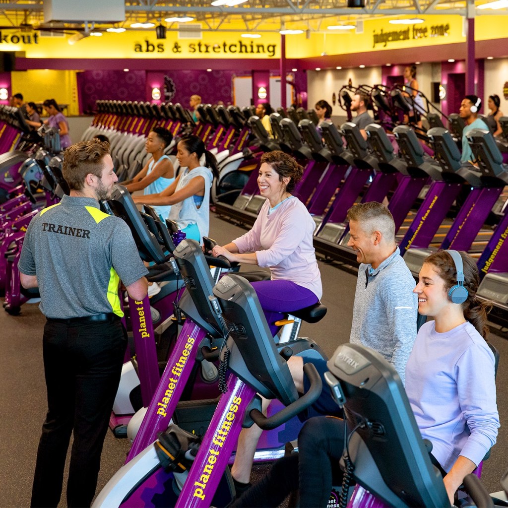 People riding stationary bikes at a planet fitness gym with a trainer standing nearby
