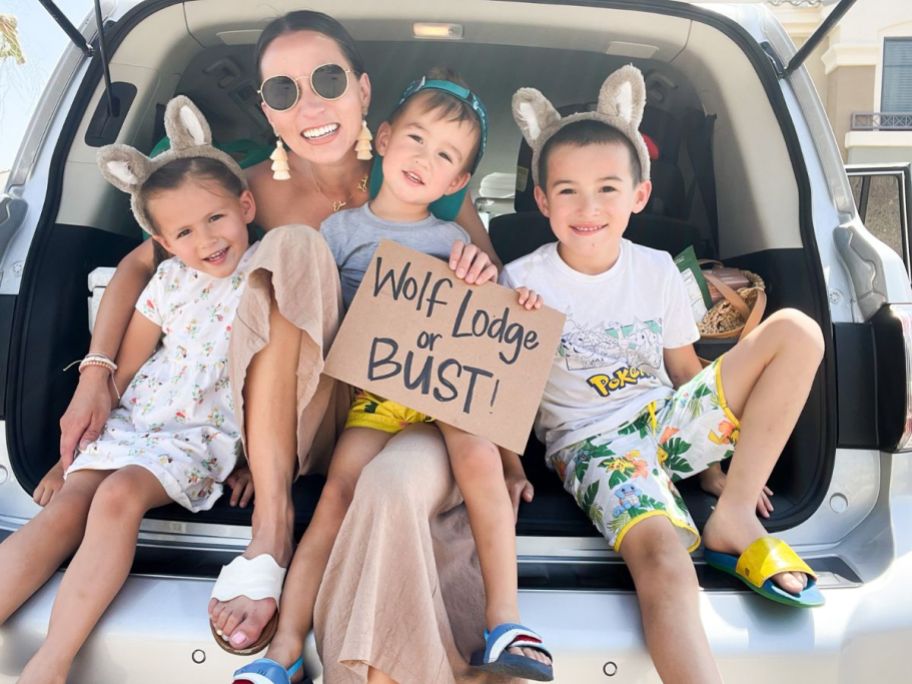 A mom and 3 kids sitting in the back of a vehicle with a Wolf Lodge or Bust! sign