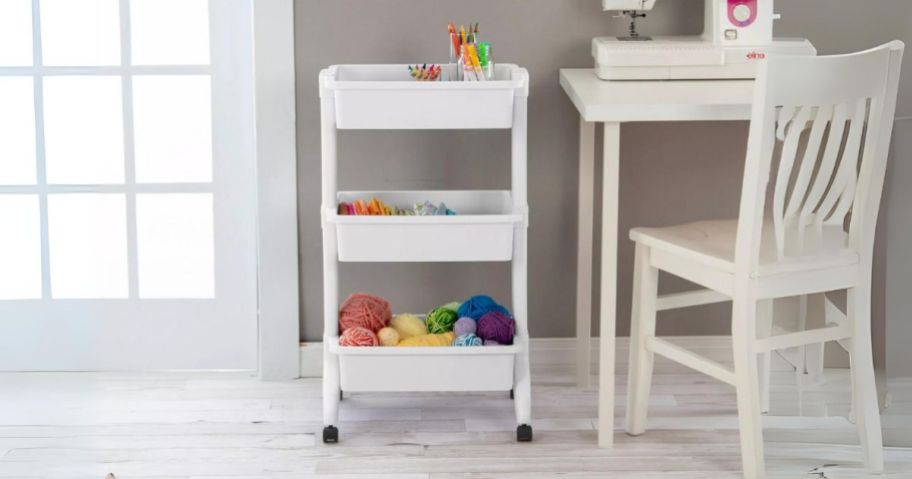 3 tier white plastic cart with craft supplies in it with a desk on one side and door on the other