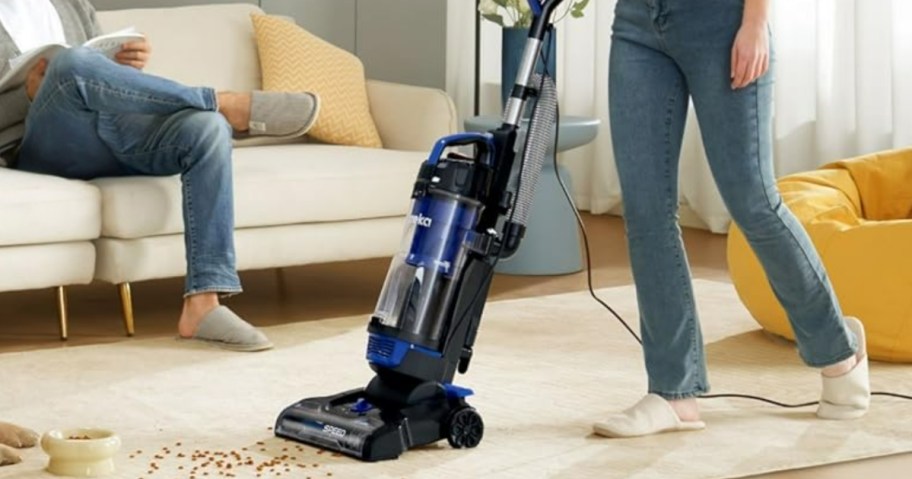 woman using a blue and black Eureka vacuum to clean up spilled dog food on carpet