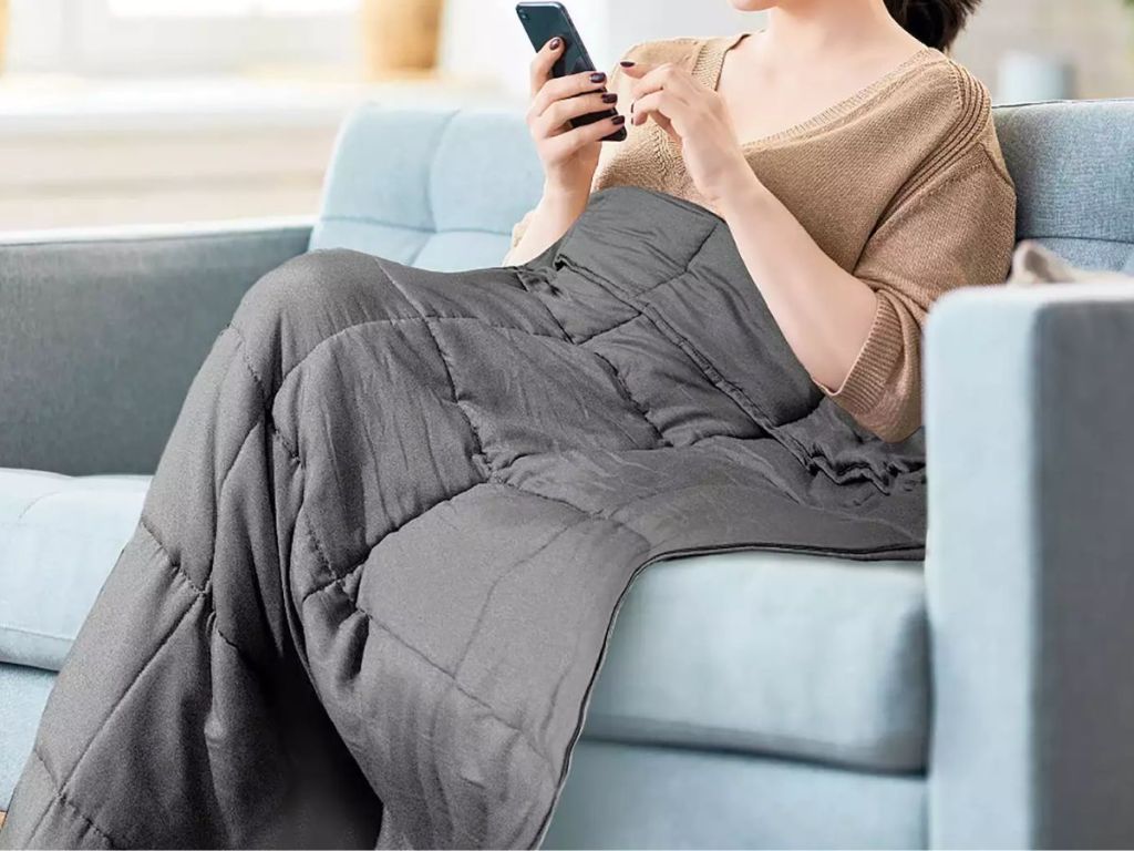 woman sitting on a couch looking at her phone covered up with a grey Microfiber Weighted Blanket