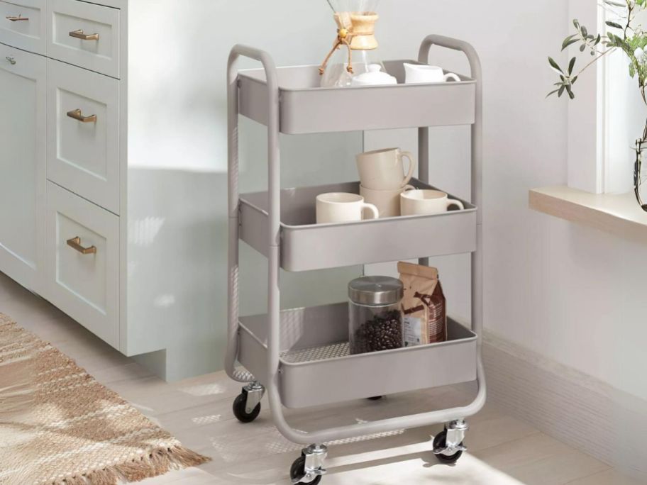 tan color metal 3 tier utility cart on wheels with kitchen items beside of a kitchen counter