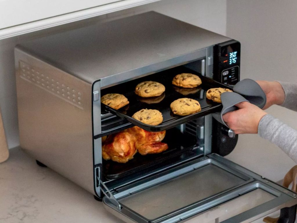 Ninja 12-in-1 Double Oven with FlexDoor shown with woman taking cookies out of top oven, chicken roasting in bottom