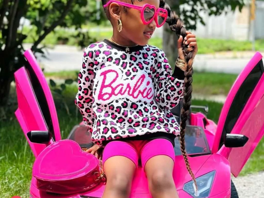 Little girl wearing a PatPat Barbie Leopard Print Shift with a pink heart shaped bag and pink heart shaped sunglasses on, sitting on a kids pink Corvette toy car