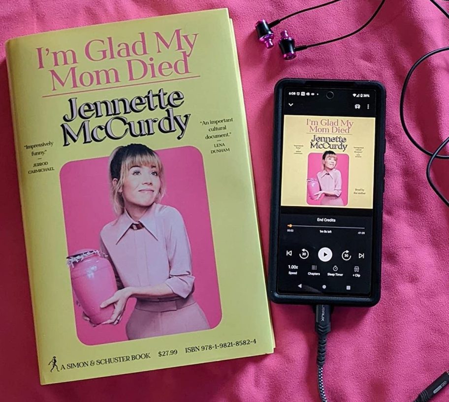 I'm glad my mom died book sitting on pink blanket next to phone with audible book on screen