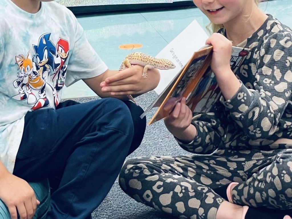 a child holding a book open with another child holding a lizard that is "reading" it