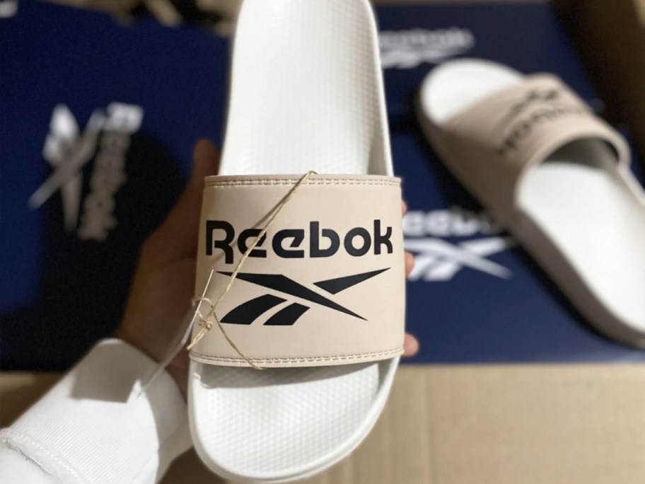 hand holding up a tan, black, and white reebok slide