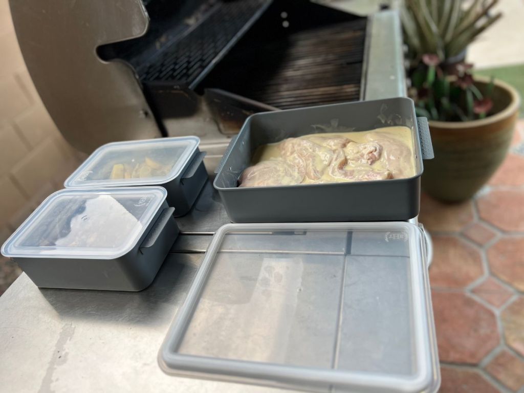 3 containers holding chicken and other food next to grill