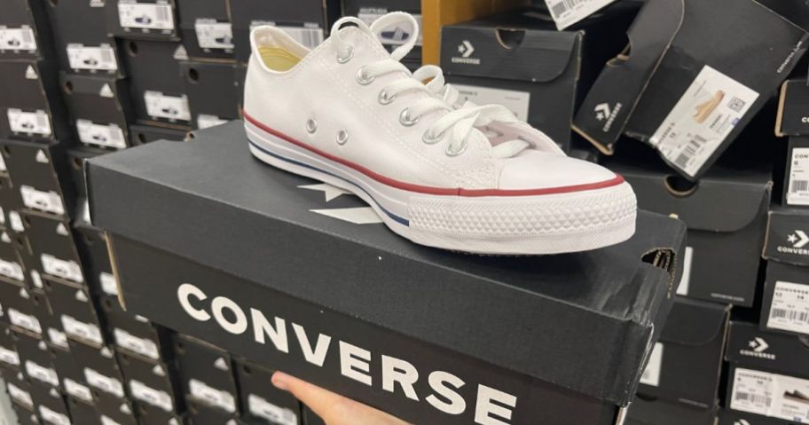 A hand holding a shoe box with a white Converse shoe perched on top