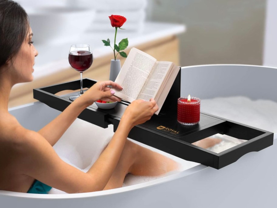 black bath tray across bathtub with woman reading book on it with candle and flowers