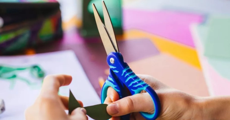 kid's hands holding a pair of blue kid's scissors and paper they are cutting