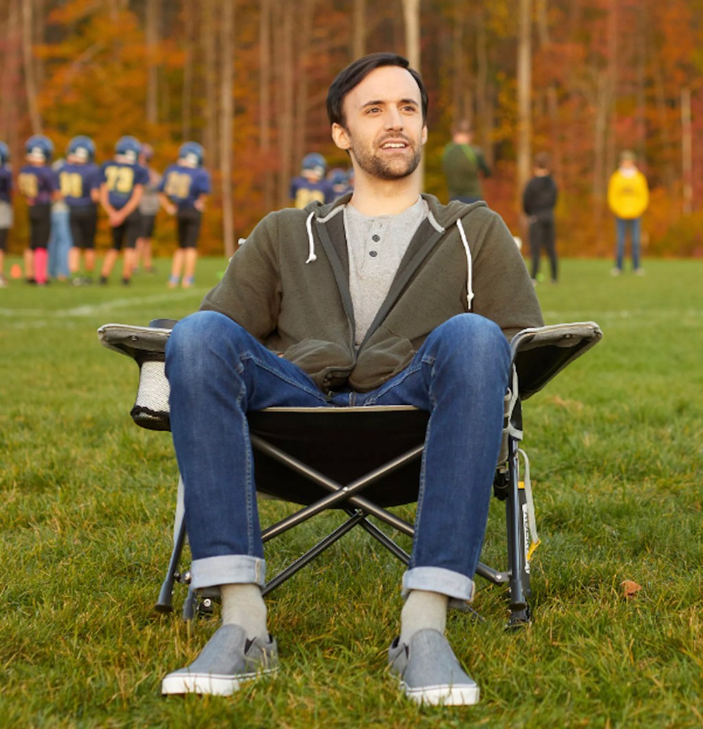 man sitting in rocker folding chair outside in grass at football game