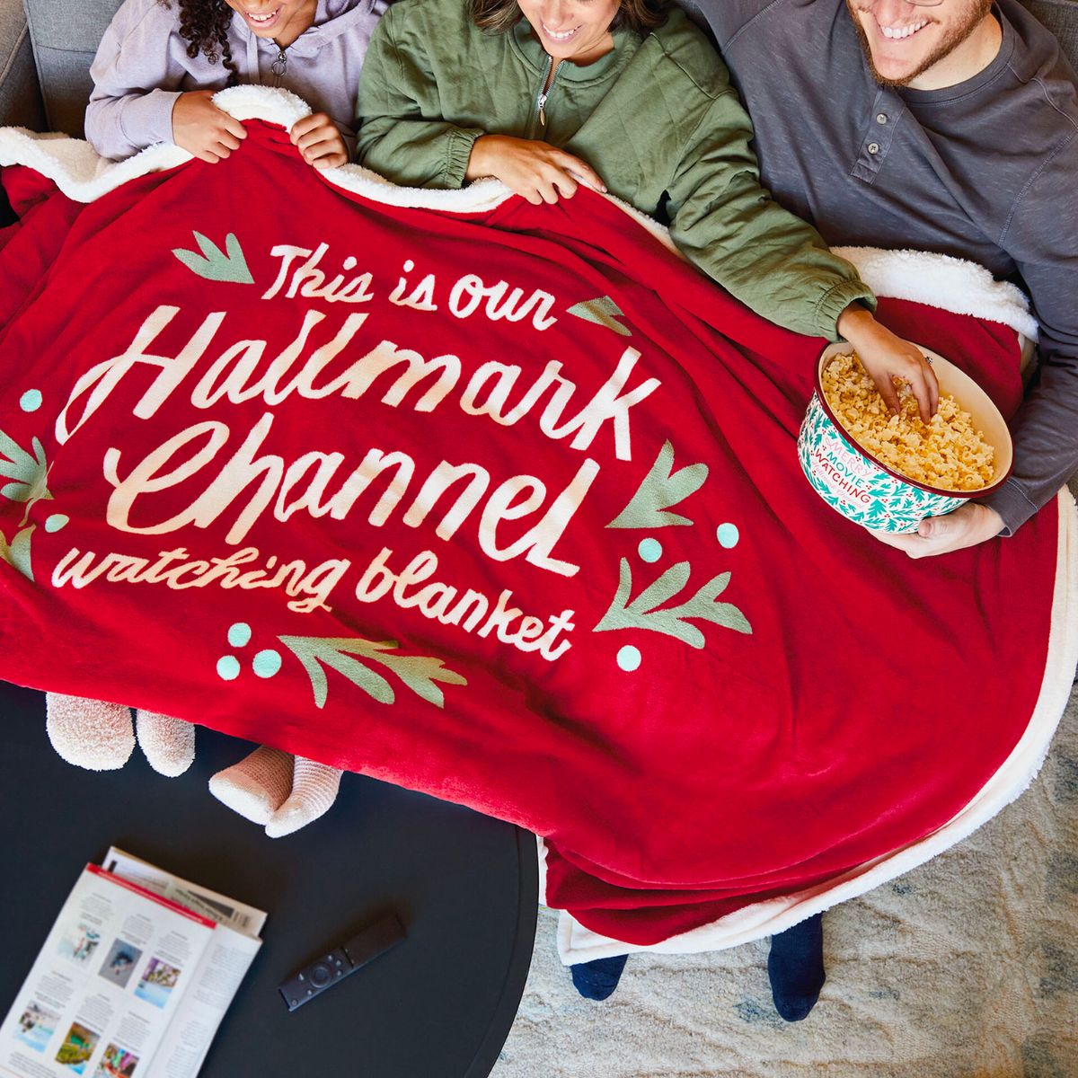 3 people snuggled under a hallmark channel oversized holiday blanket eating popcorn