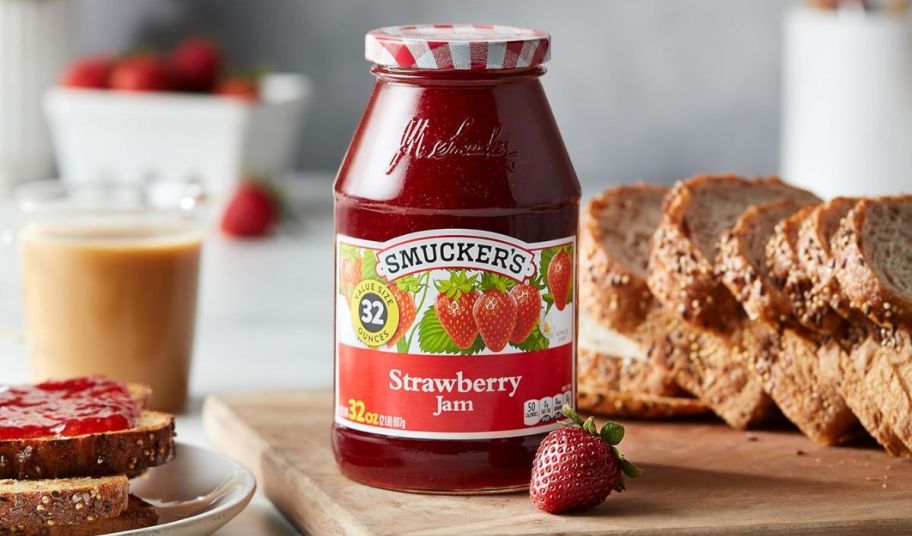 a 32 oz jar of Smuckers strawberry preserves on a wooden cutting board next to a fresh strawberry