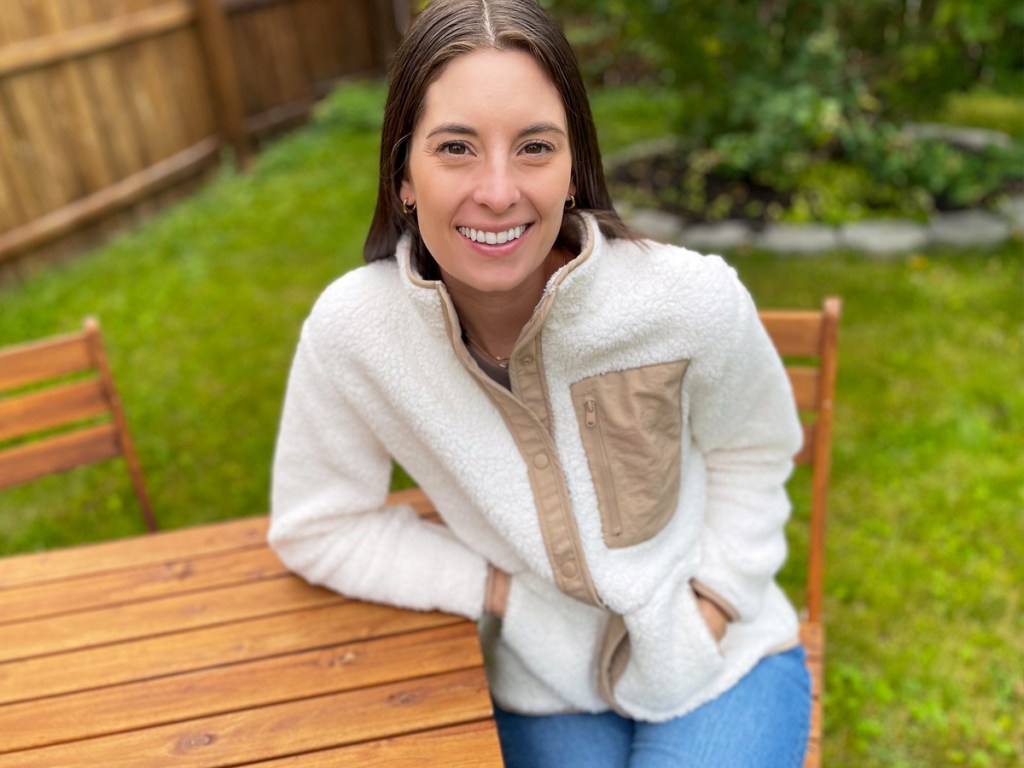 woman sitting outside at wood table wearing a white sherpa jacket and jeans
