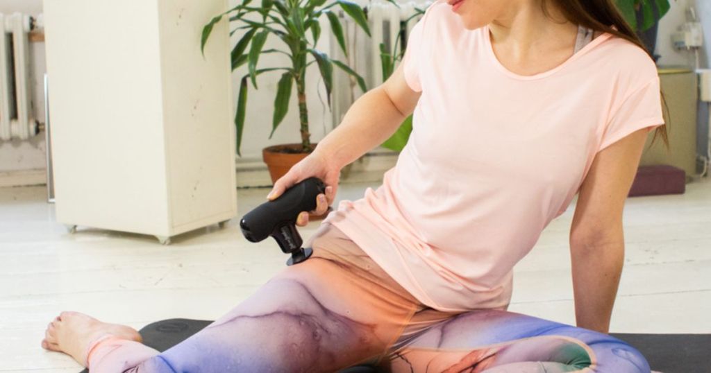 Woman using a BOB AND BRAD Q2 Mini Pocket-Sized Deep Tissue, Portable Percussion Muscle Massager Gun on her hip while sitting on a yoga mat