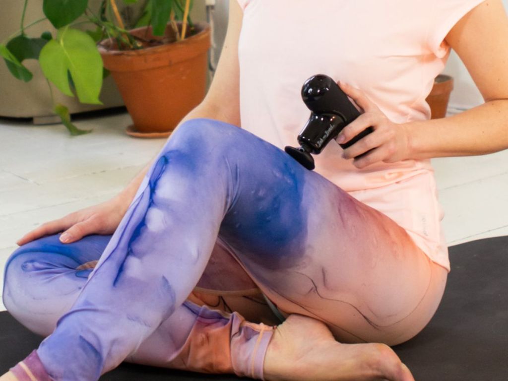 Woman sitting on a yoga mat using a BOB AND BRAD Q2 Mini Pocket-Sized Deep Tissue, Portable Percussion Muscle Massager Gun on her leg