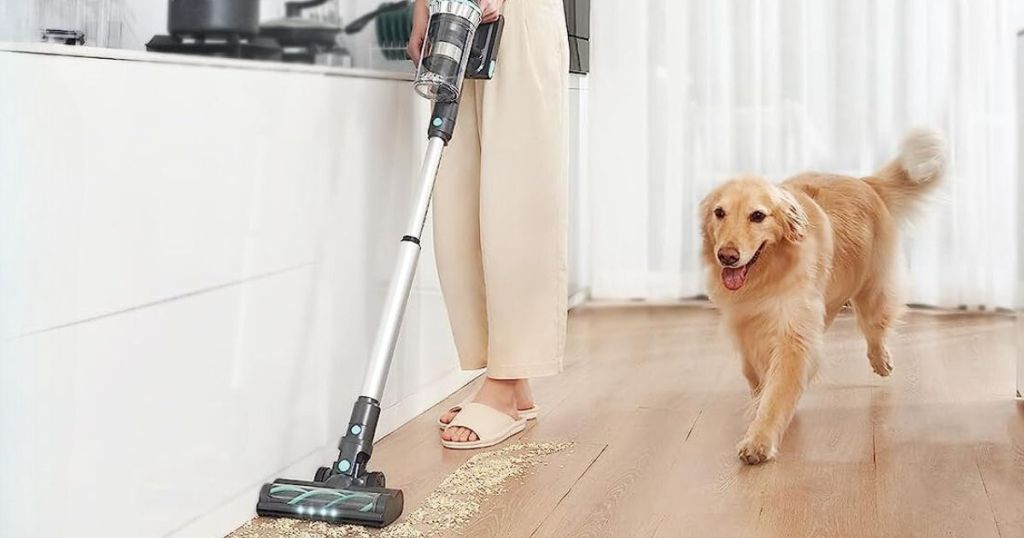 person vacuuming floor with stick vacuum and dog running beside it