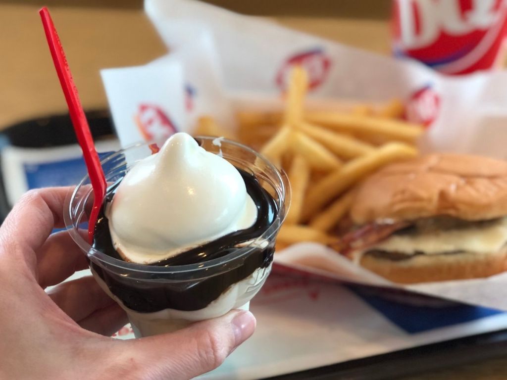 A hand holding a Dairy Queen ice cream sundae in front of a basket with a burger and fries
