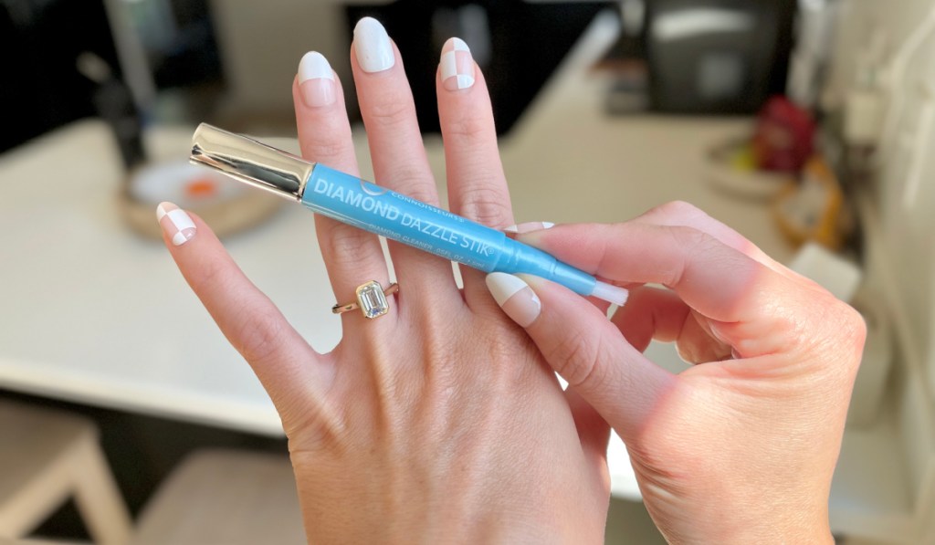 A hand holding a Diamond Dazzle stick jewelry cleaner next to a sparkling engagement ring