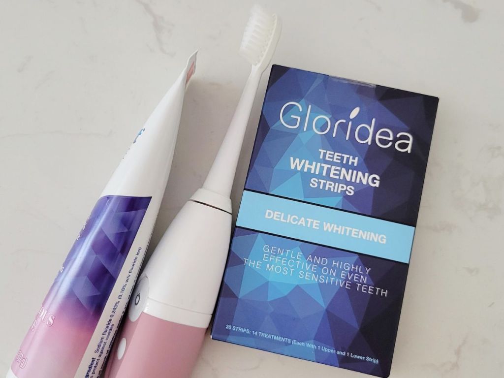 Gloridea Teeth Whitening Strips next to an electric toothbrush and a tube of toothpaste