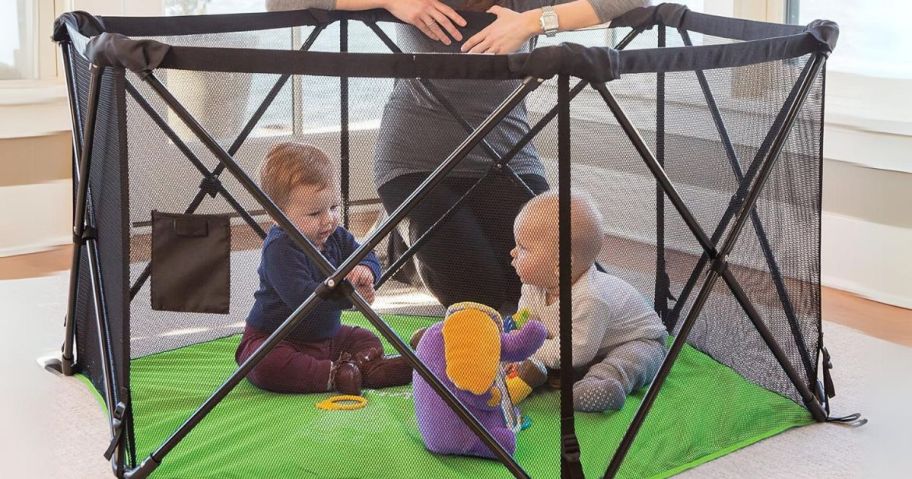 woman looking over a pop up play pen with a green floor that a baby and toddler are playing in with toys