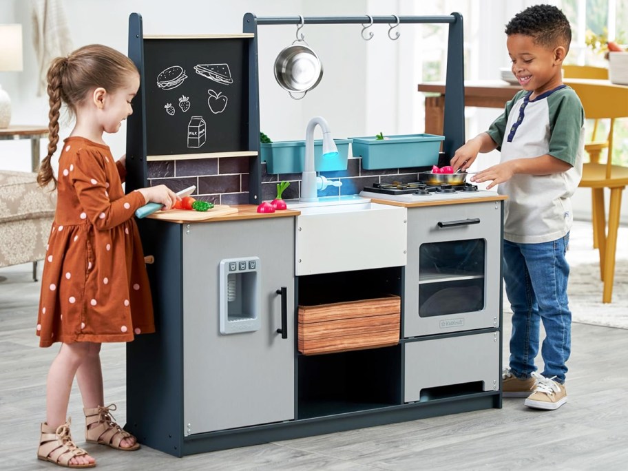 two kids standing on either side of a kidkraft wooden kitchen playset