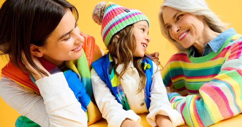 A woman, child and older woman sitting at a table, looking at each other lovingly while wearing clothing and accessories from the Crayola X Kohl's collection.