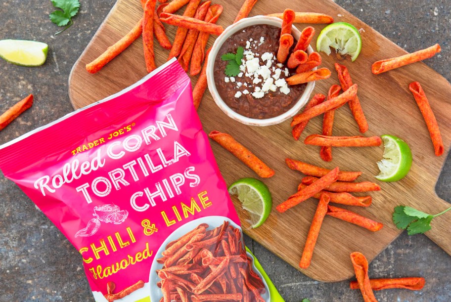 Trader Joe's Rolled Corn Tortilla Chips Chili and Lime scattered on wood board next to bean dip and cotija cheese garnish with lime wedges and cilantro