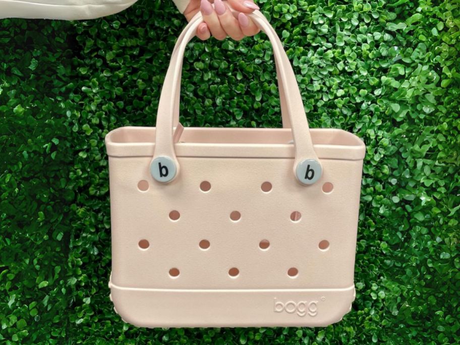 hand holding a light pinkish peach color Bitty Bogg Tote in front of a greenery background