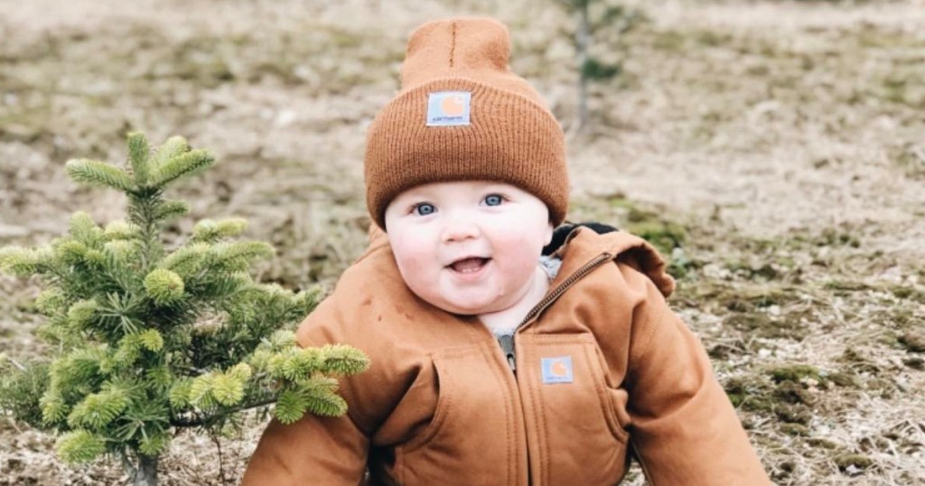 baby wearing brown carhartt hat next to small tree