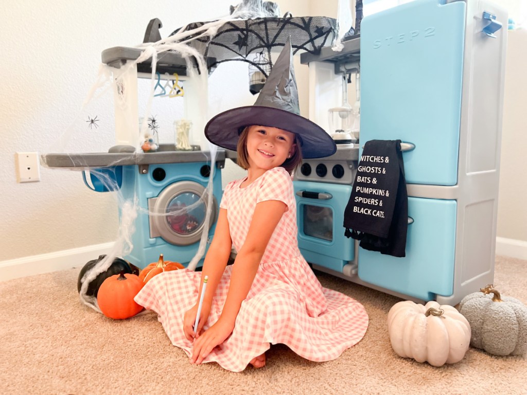 girl in witch hat next to play kitchen
