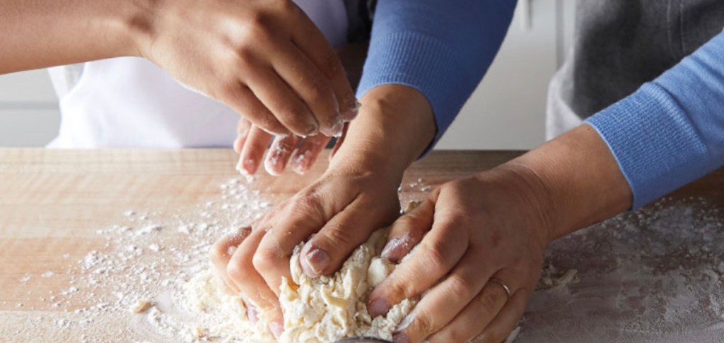 two peoples hands kneading pizza dough on butcher block countertop